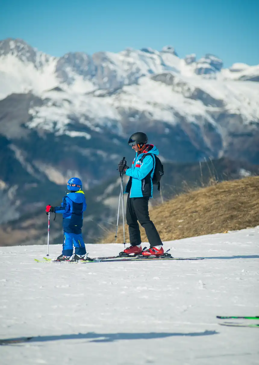 Adulto e criança esquiando numa encosta nevada, com montanhas ao fundo